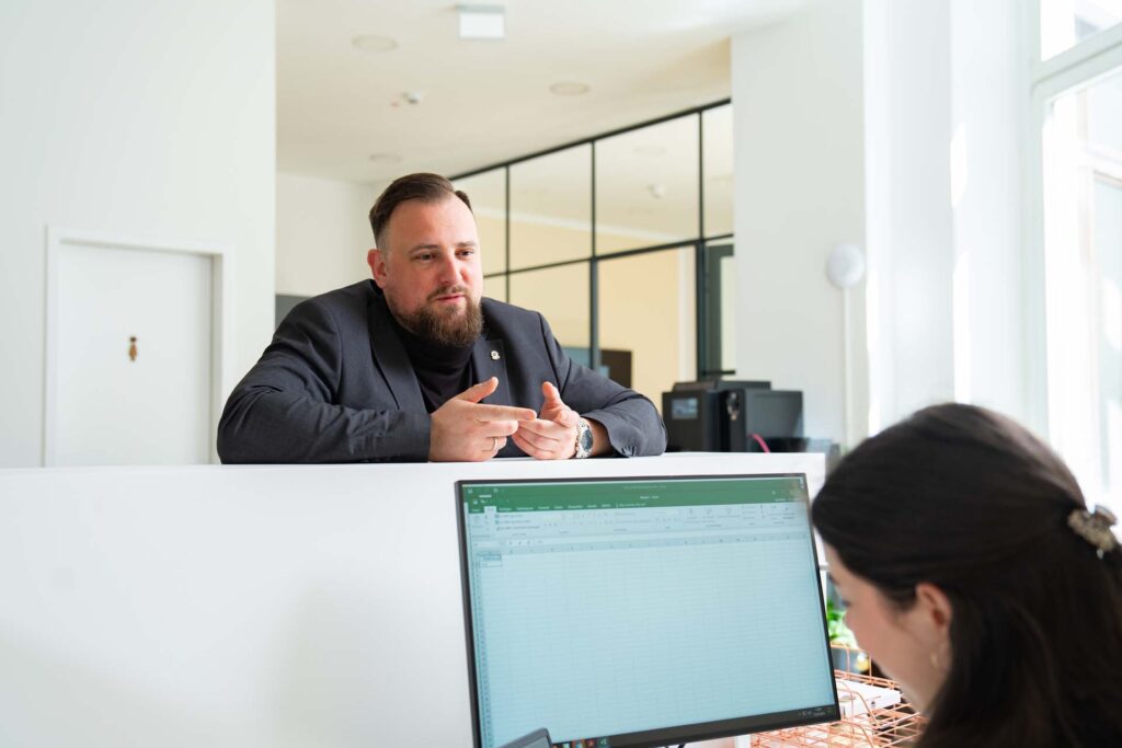 Ein Mann im Anzug lehnt an einem weißen Tresen und spricht mit einer Frau, die in einem Büro mit großen Fenstern und moderner Einrichtung an einem Computer mit einer Tabellenkalkulation arbeitet, über Interim Management Consulting.