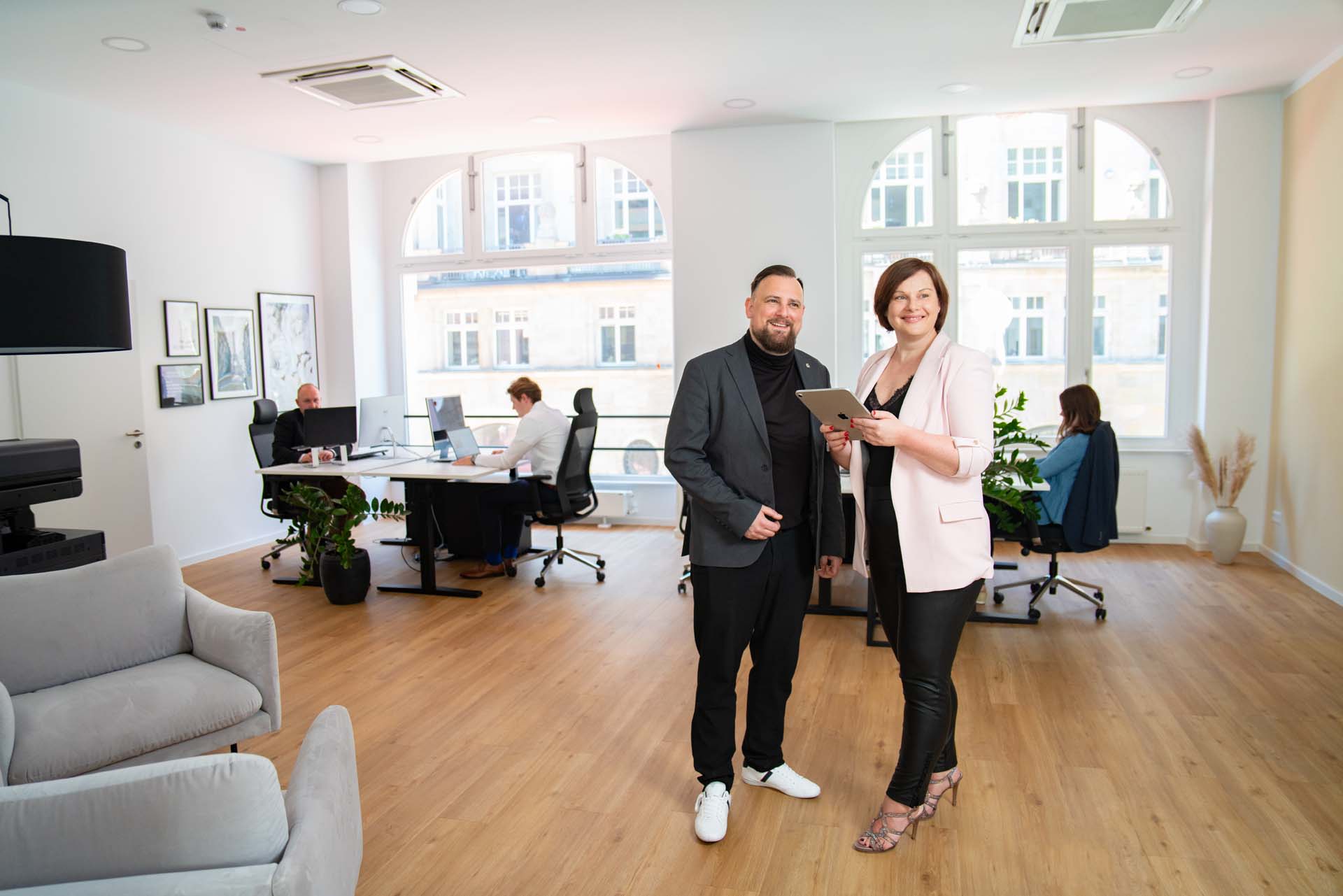 Zwei Personen stehen lächelnd im Vordergrund eines hellen, modernen Büros in Dresden und verweisen auf unsere Kompetenz in der existenzgründung steuerberatung; im Hintergrund arbeiten drei Kollegen an Schreibtischen.